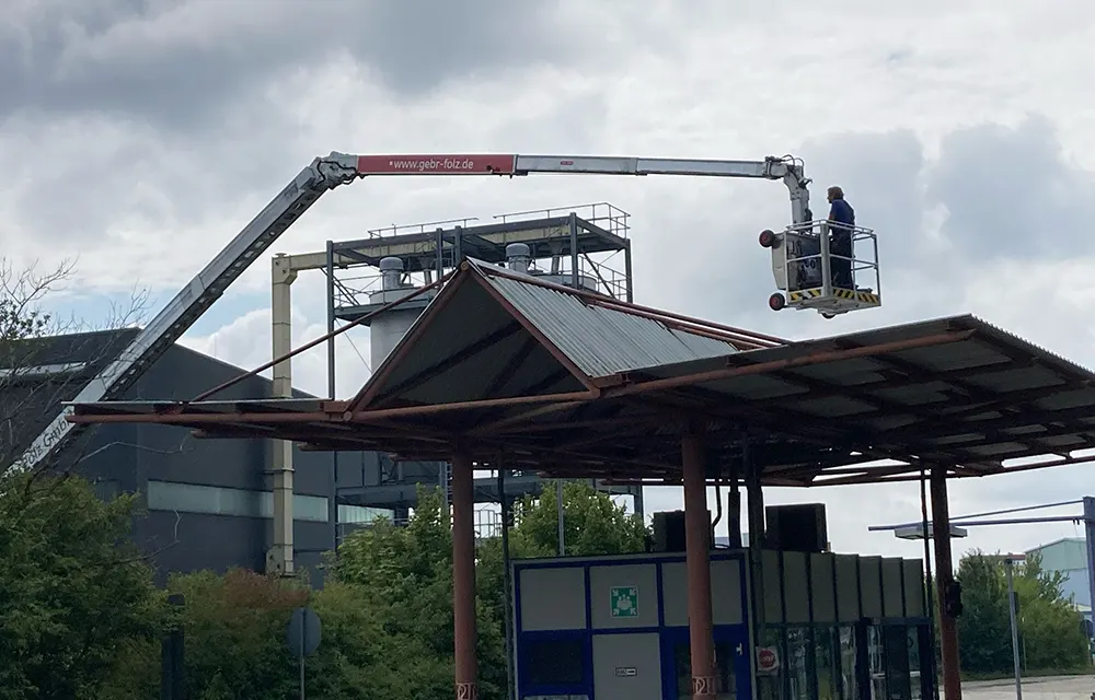 Arbeiter steht im Korb einer Hebebühne über Vordach, Industriegebäude im Hintergrund.
