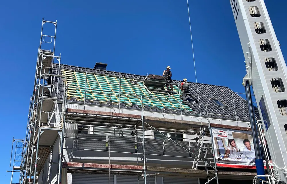 Zwei Dachdecker arbeiten auf einem eingedeckten Steildach mit Gerüst und Sicherheitsausrüstung.