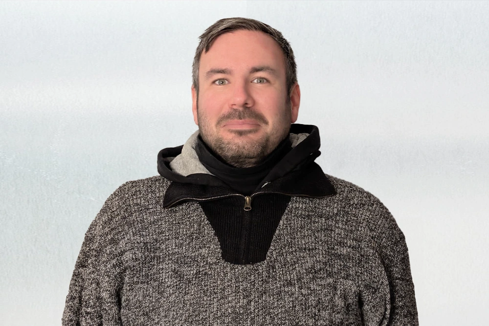 Porträt von Mark mit kurzen dunklen Haaren und Bart, der vor hellem Hintergrund in die Kamera blickt und einen grauen Pullover trägt.