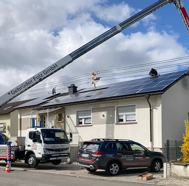 Kran hebt Arbeiter zum Dach, Montage von Taubenschutz an PV-Anlage auf Einfamilienhaus bei sonnigem Wetter.