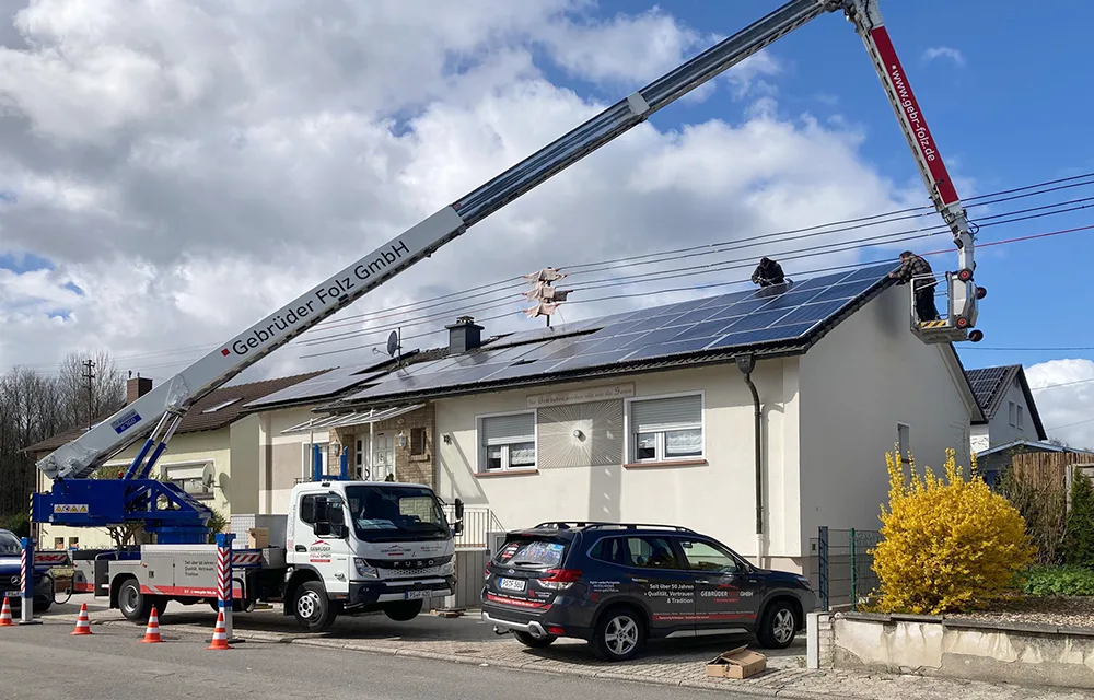 Kran hebt Arbeiter zum Dach, Montage von Taubenschutz an PV-Anlage auf Einfamilienhaus bei sonnigem Wetter.